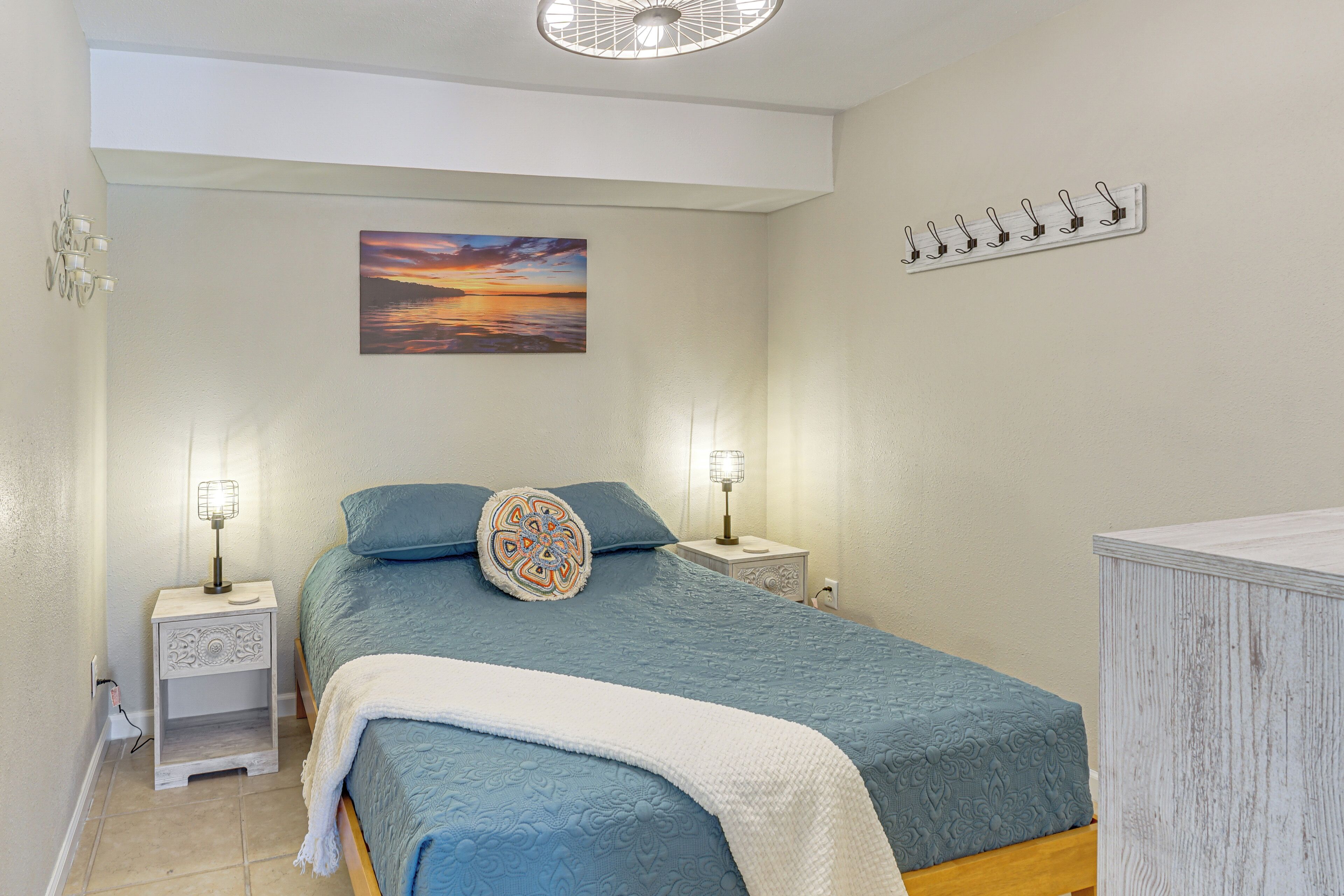 Un dormitorio con cama, lámparas de noche, una lámpara de pared, un cuadro y un perchero.