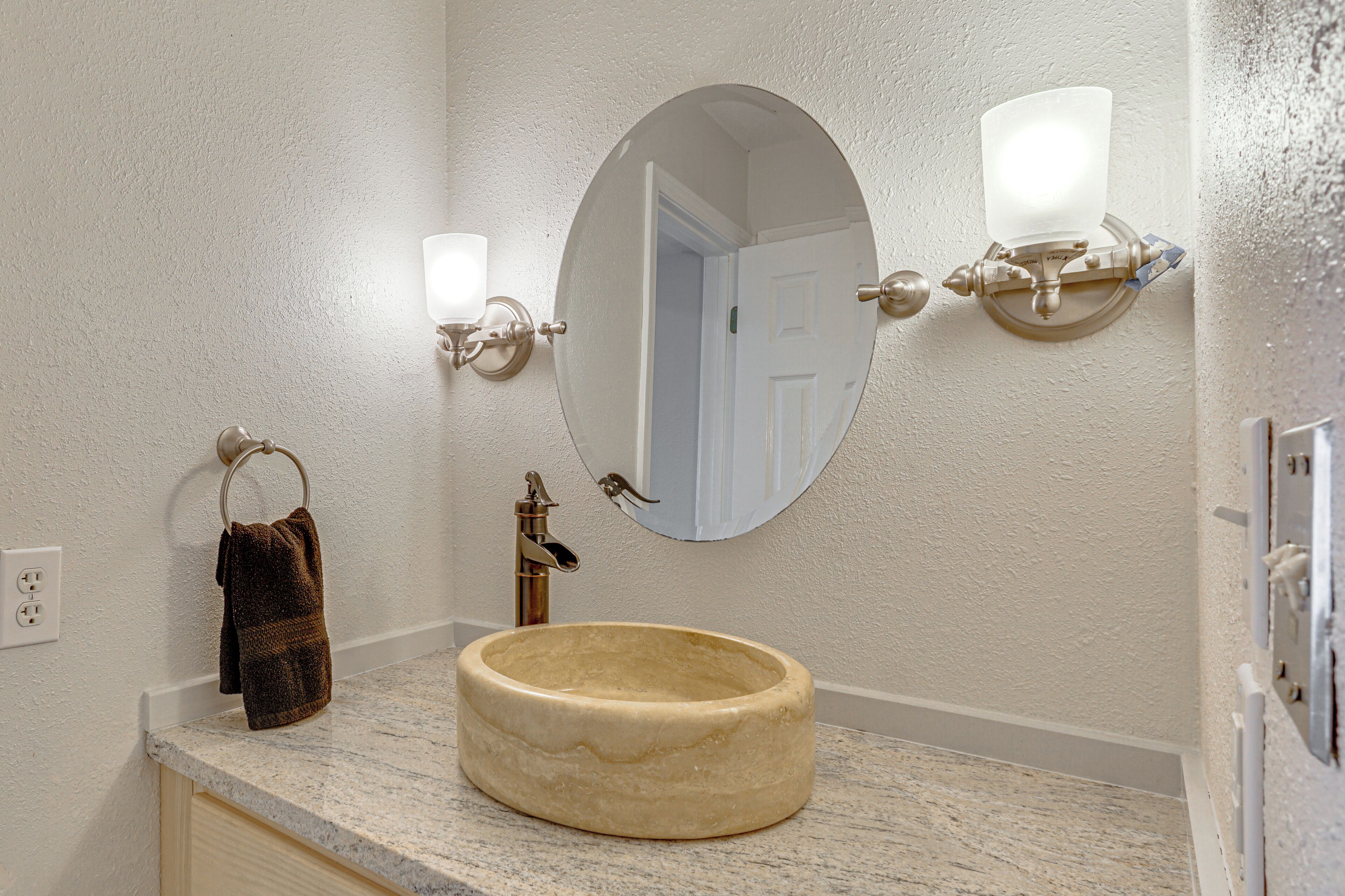 A bathroom with a round marble sink, a towel ring, a mirror with wall sconces, and a door in the background.