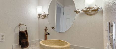 A bathroom with a round marble sink, a towel ring, a mirror with wall sconces, and a door in the background.