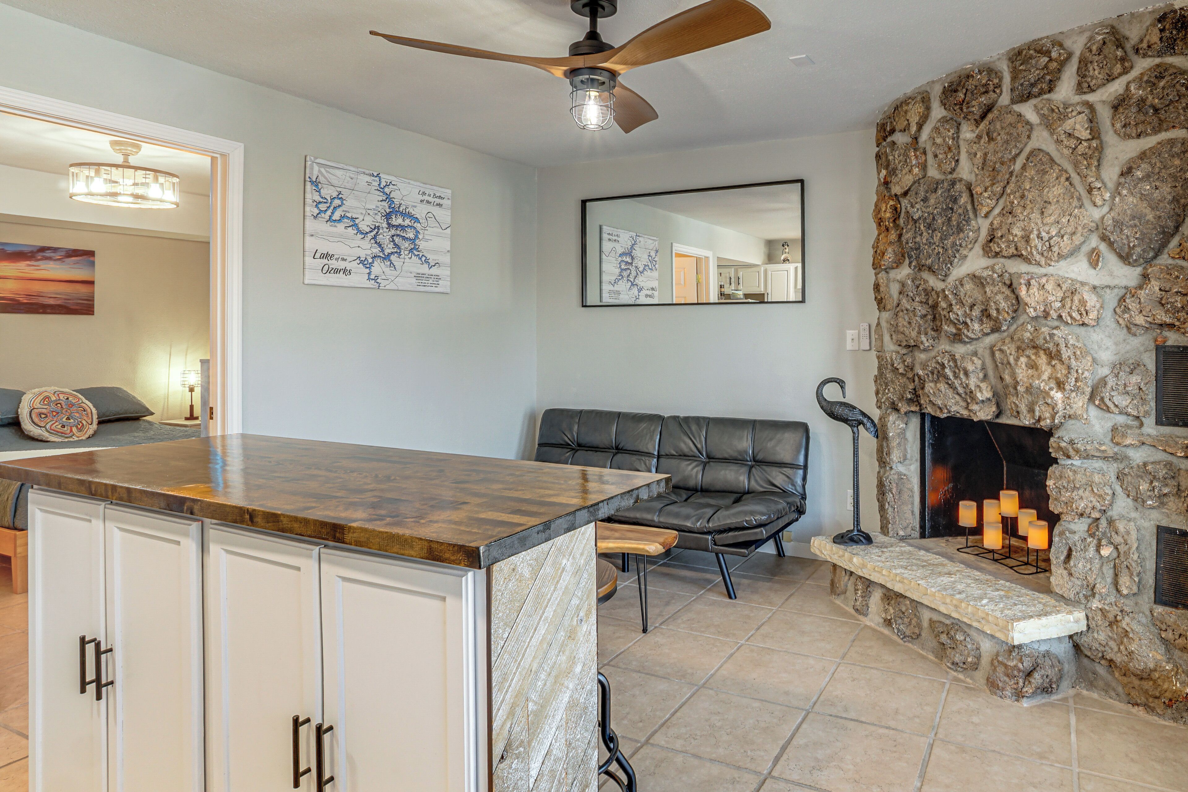 A living room with a stone fireplace, a ceiling fan, and a kitchen area with a wooden countertop.