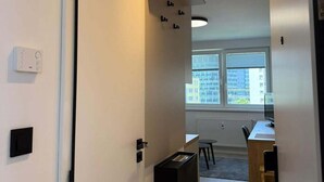 A modern interior with a door, a wall-mounted light fixture, a window with blinds, and a desk with a chair. - Beautiful 1-bedroom apartment in cozy Košický kraj perfect for relaxing (Košický kraj)