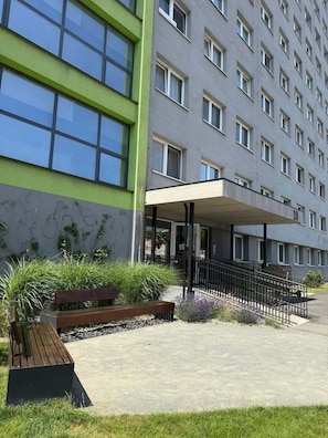 A multi-story building with a green facade, a covered entrance, and a paved area with a bench and plants.