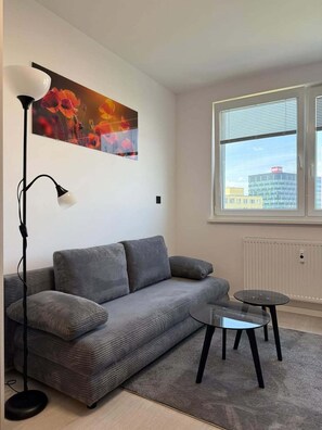A modern living room with a grey sofa, a floor lamp, a small round table, and a window with a city view.