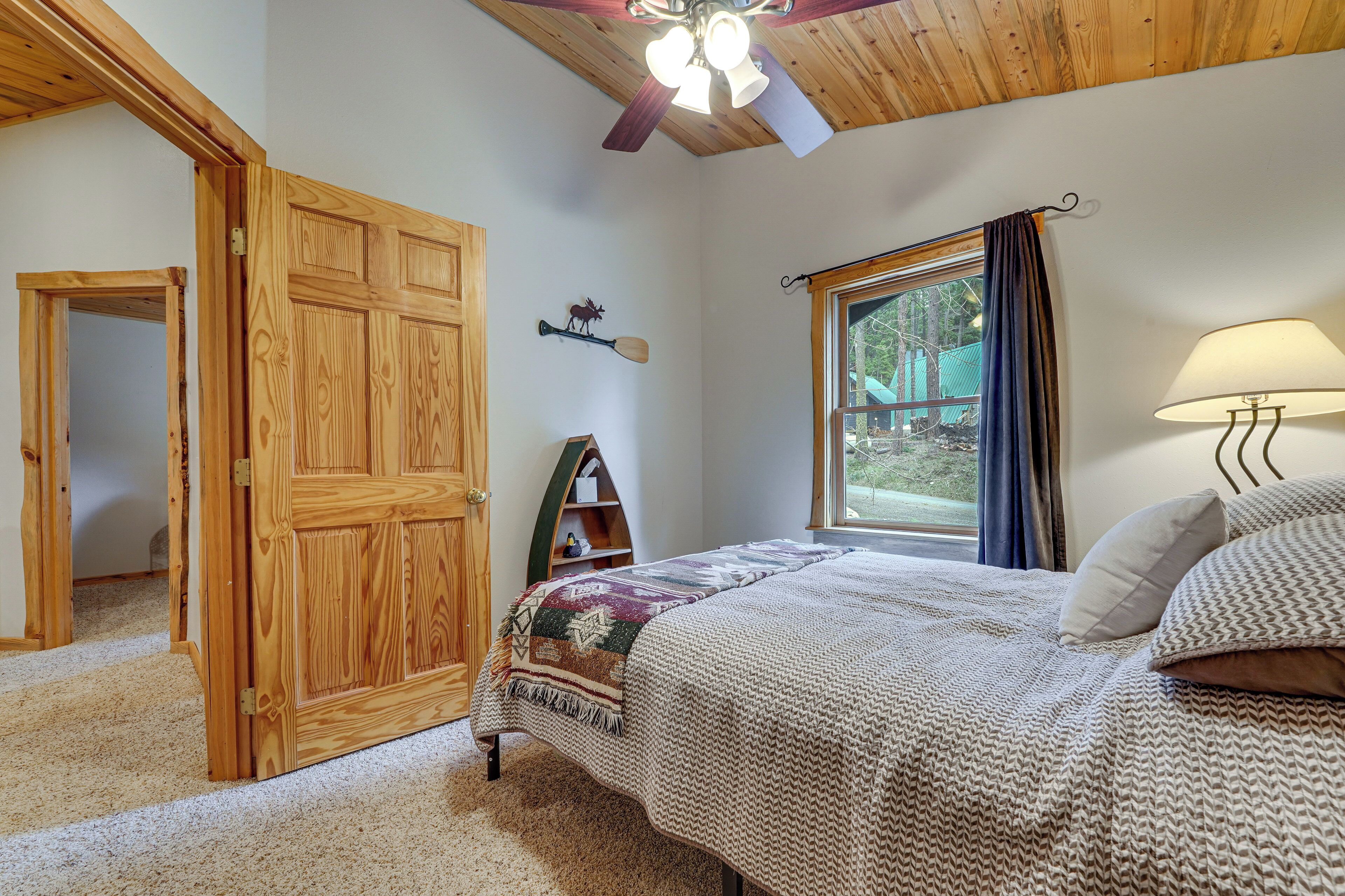 A bedroom with a wooden door, a bed with a patterned blanket, a nightstand, a lamp, and a window with a view of the outdoors.