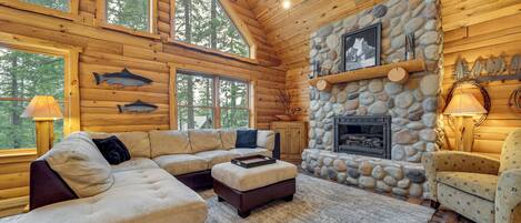 A log cabin interior with a stone fireplace, wooden walls, and a large window overlooking trees.