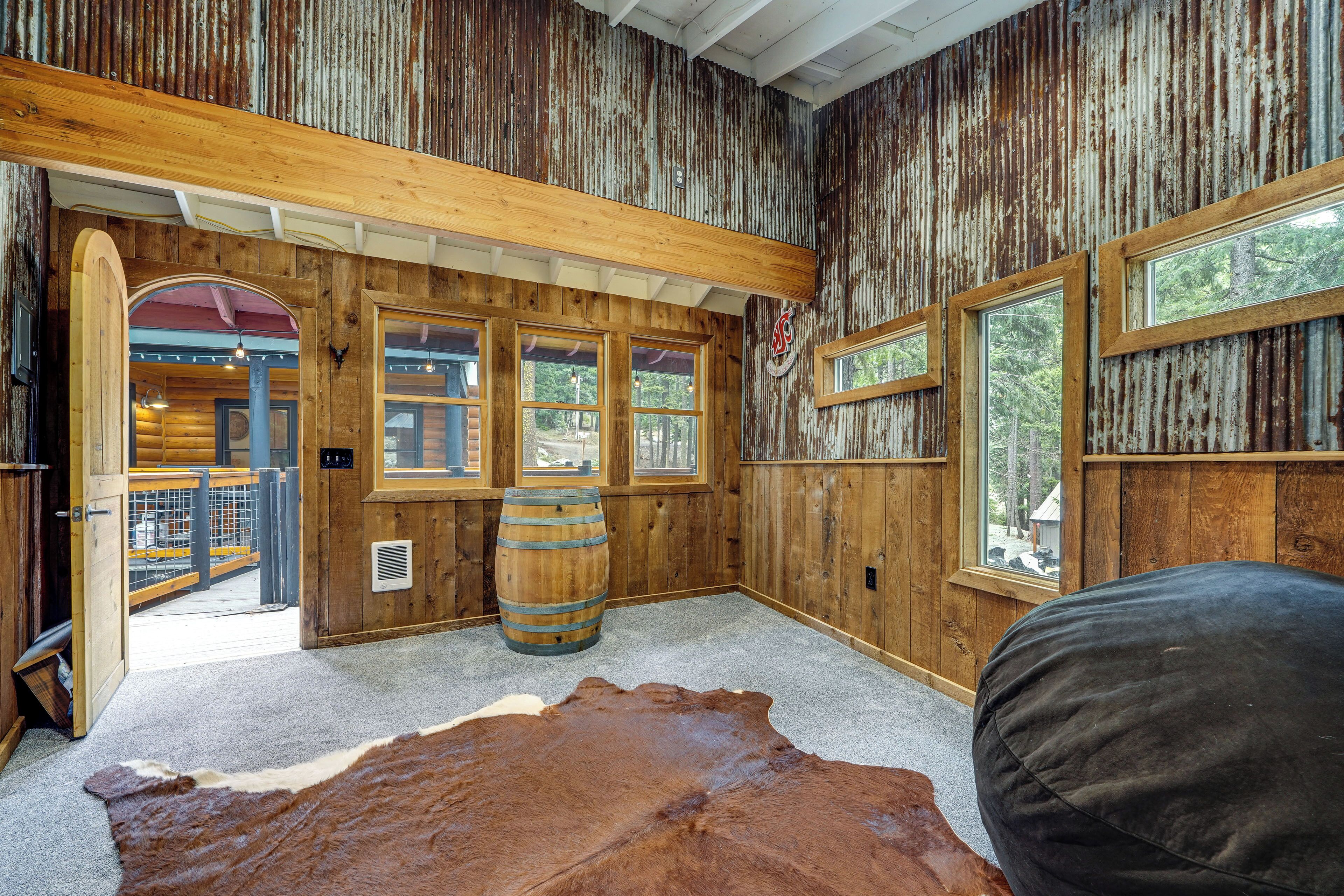 A rustic interior with wooden walls, large windows, a wooden barrel, and a leather rug.