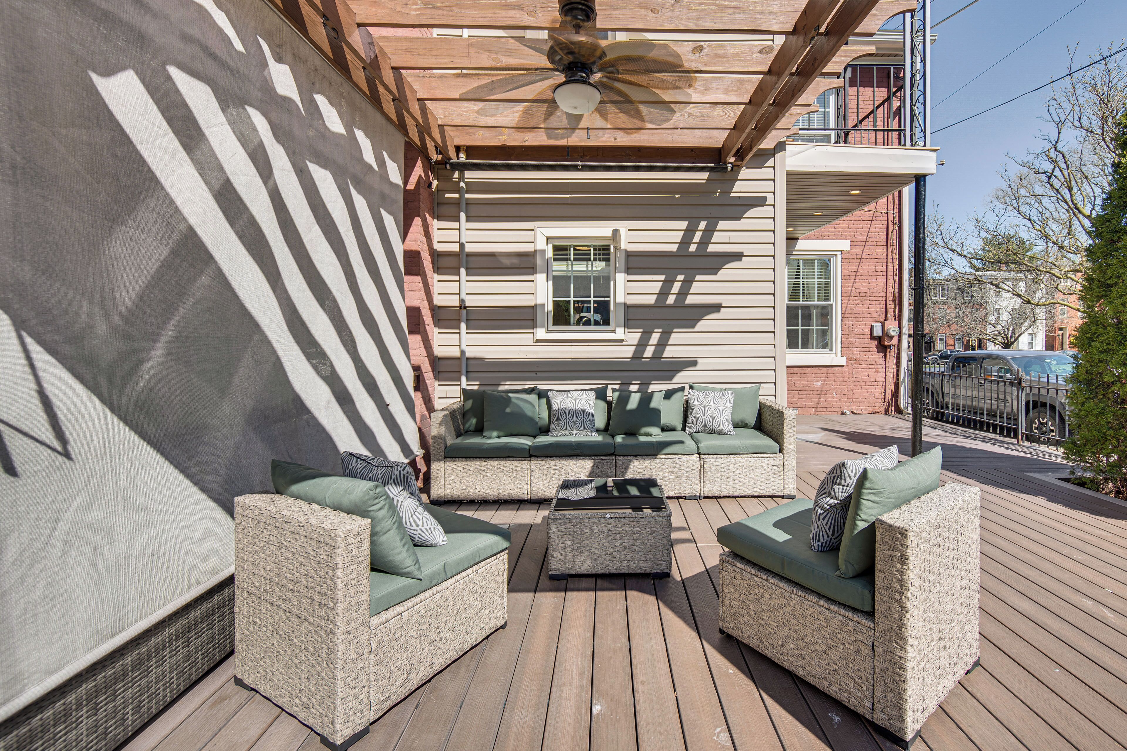 Eine Terrasse im Freien mit Korbmöbeln, einem Deckenventilator und Blick auf ein Backsteinhaus.