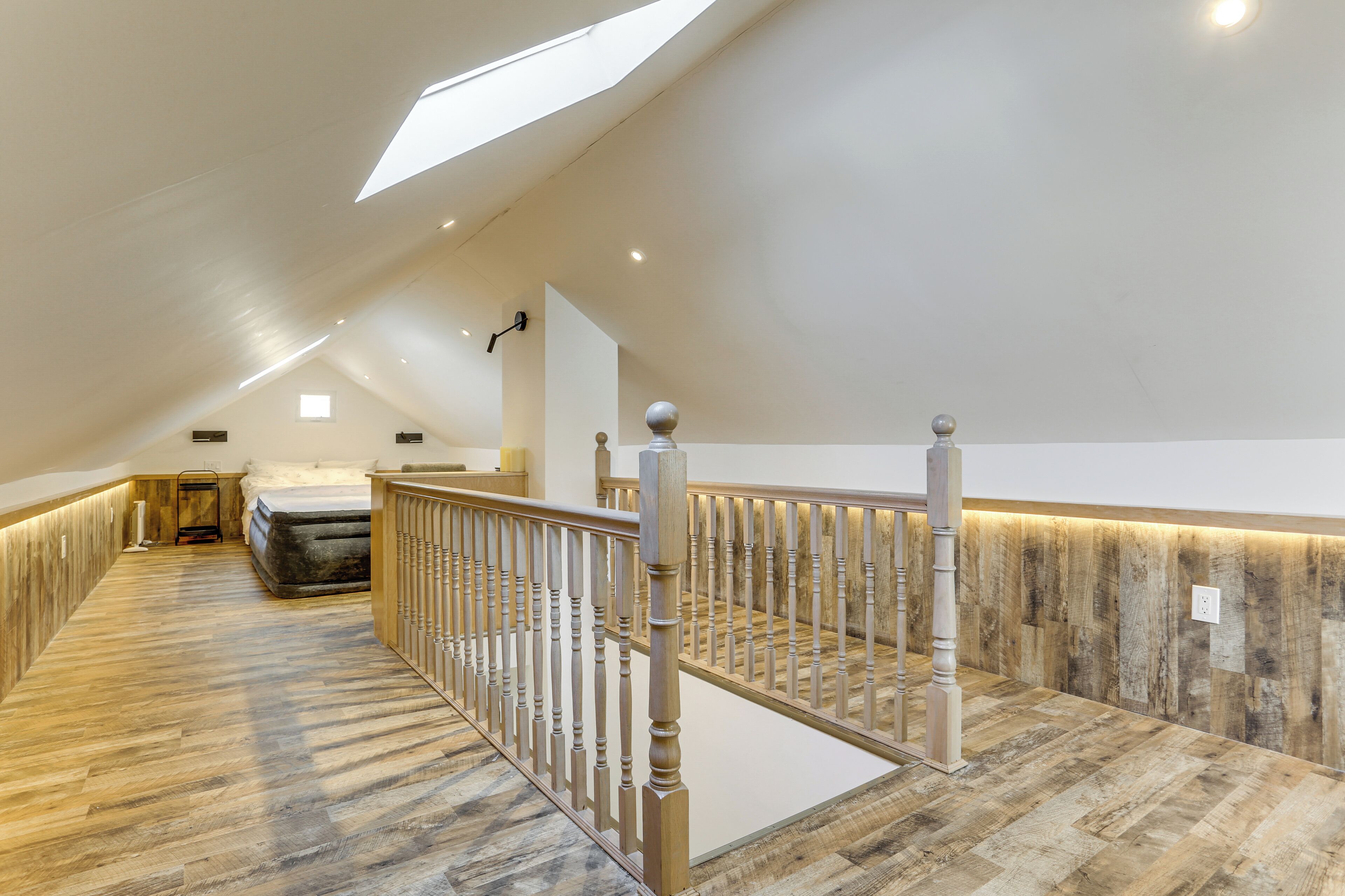 A spacious attic bedroom with a sloped ceiling, a wooden floor, a bed, and a staircase with a wooden railing.