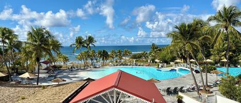 A resort pool area with a view of the ocean, palm trees, and a covered seating area.
