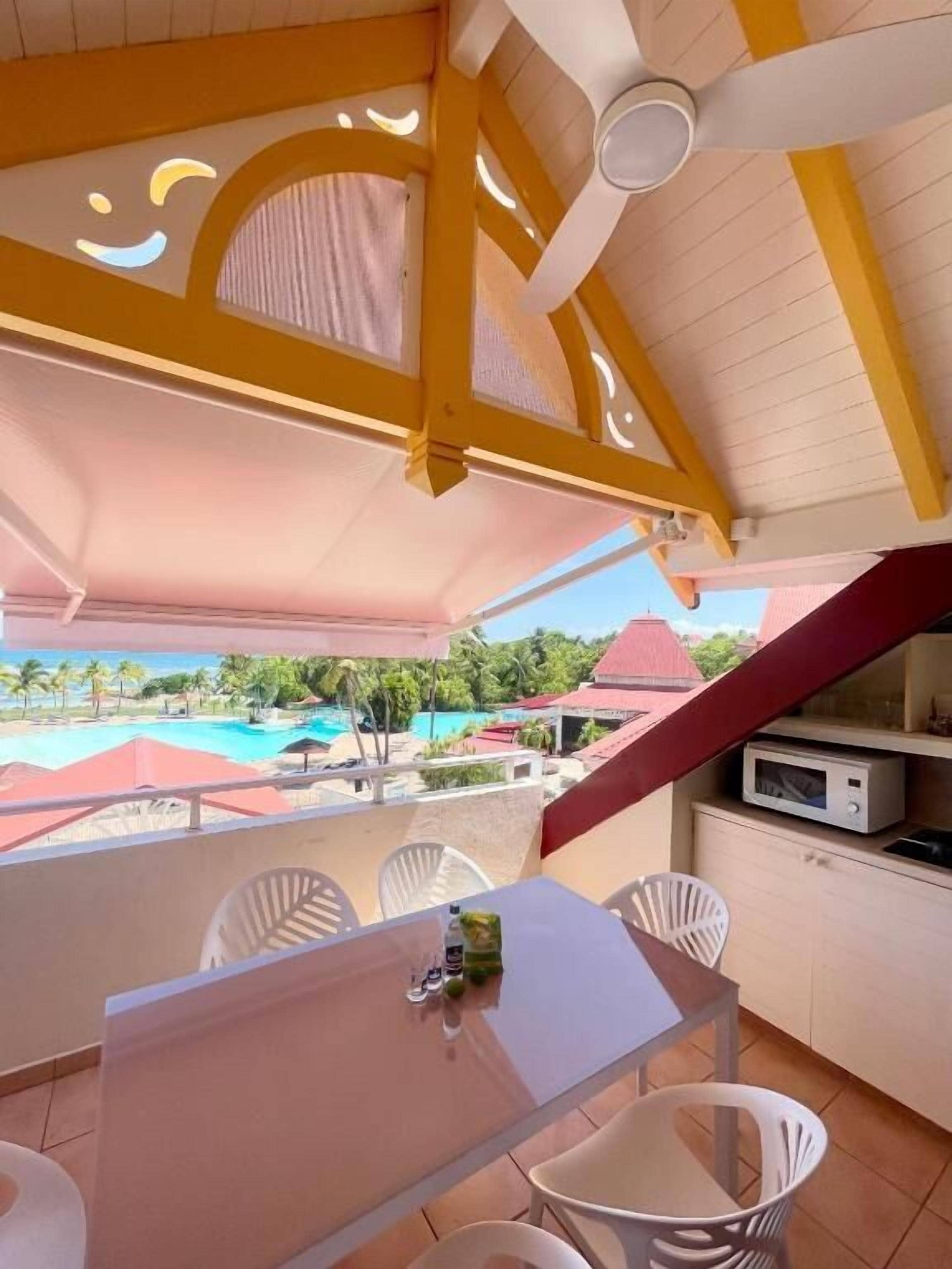 Een balkon met een tafel en stoelen, een plafondventilator en uitzicht op een zwembad en resortgebouwen.
