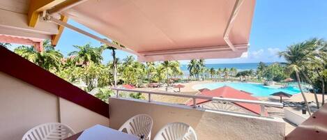 A balcony with a table, chairs, and a view of a pool and beach.