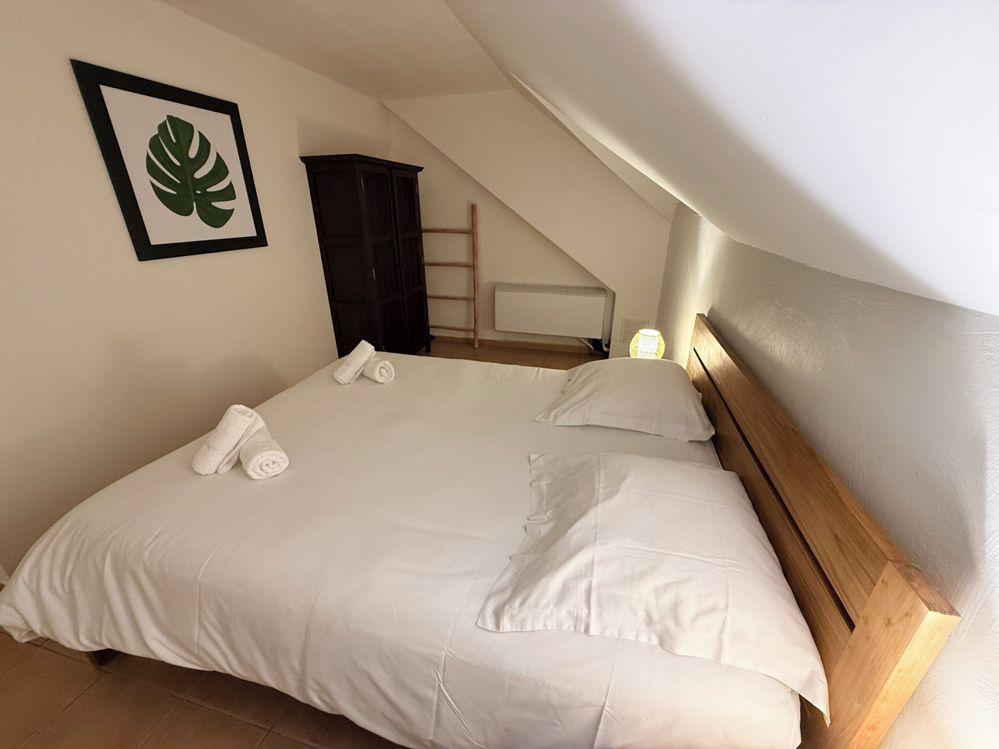 A neatly made bed with white linens and towels, a framed leaf artwork on the wall, and a wooden headboard.