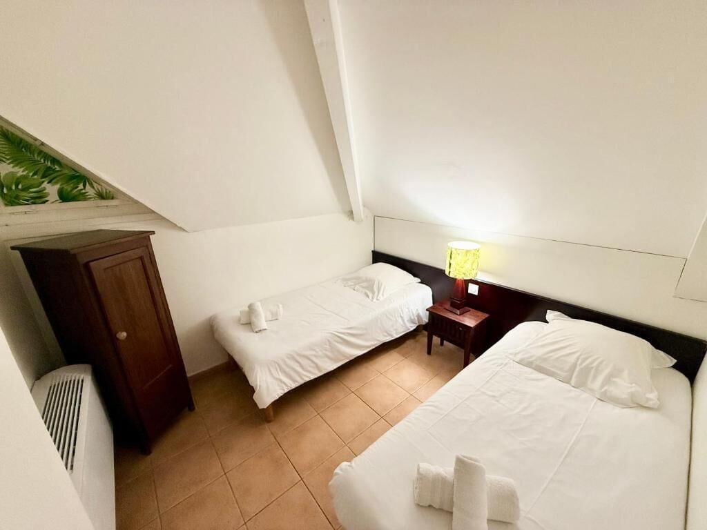 A hotel room with two beds, a nightstand with a lamp, a wooden wardrobe, and a radiator.