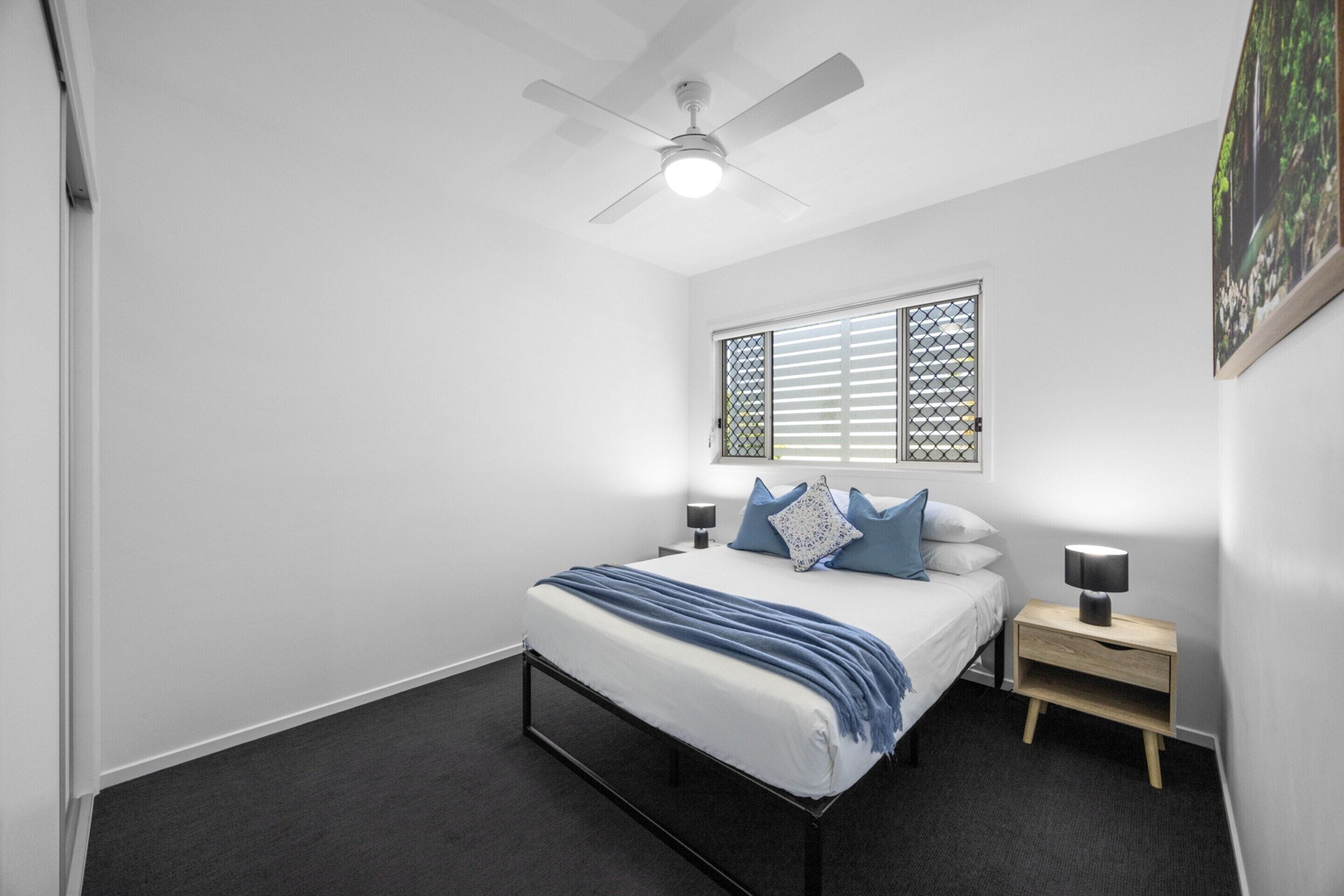 A bedroom with a bed, a nightstand, a ceiling fan, a window with blinds, and a wall-mounted picture.