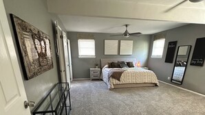 Un dormitorio con cama, mesitas de noche, cómoda, ventilador de techo y obras de arte en las paredes.