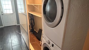 Un lavadero con lavadora y secadora de carga frontal, estantes de madera y piso de baldosas.