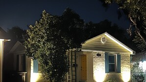 Una casa residencial de noche con iluminación exterior, una puerta principal y un pequeño jardín delantero.