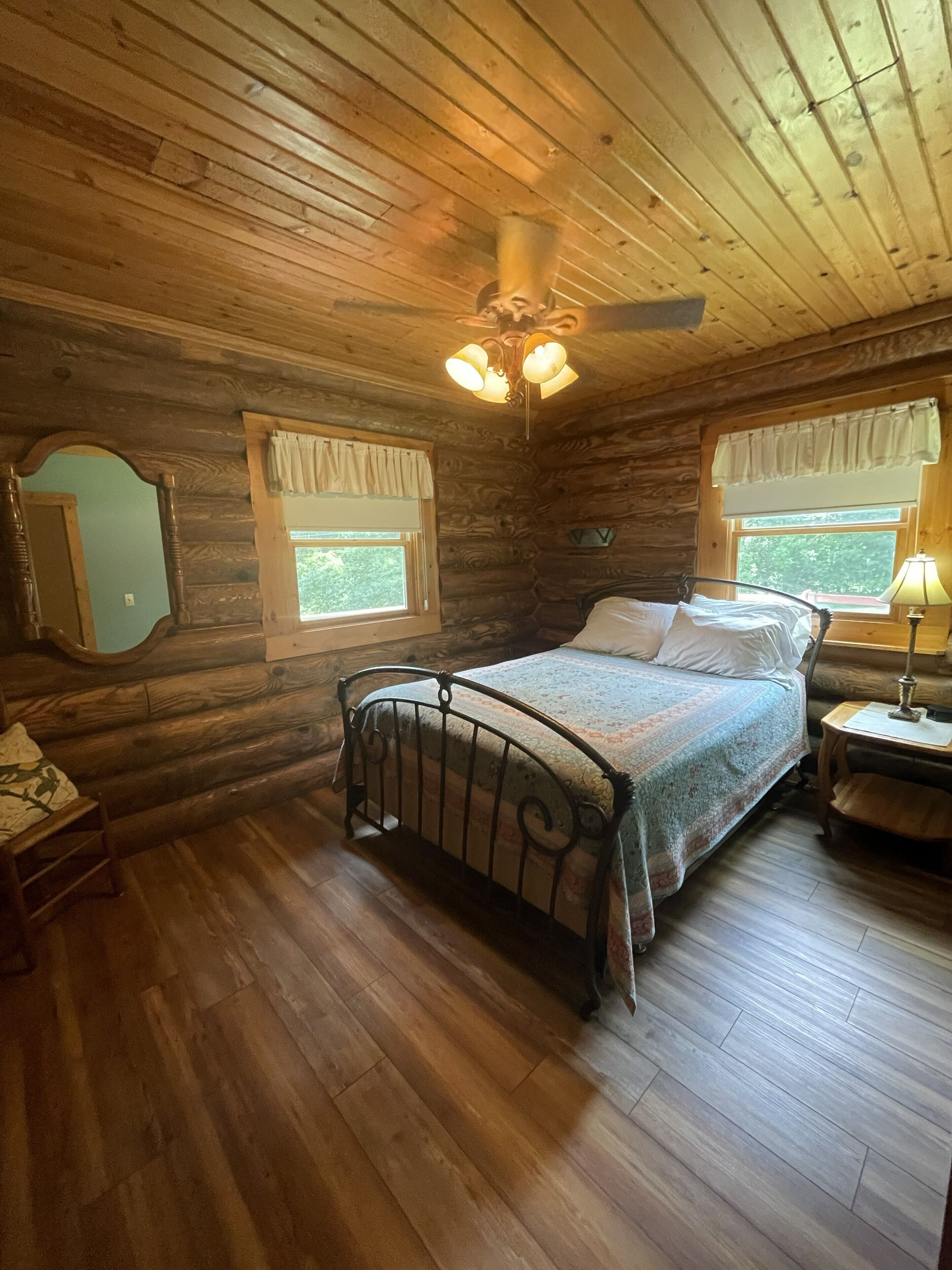 Camera da letto in una baita di legno, con un letto, un comodino, una lampada, una ventola a soffitto e una finestra con tende.