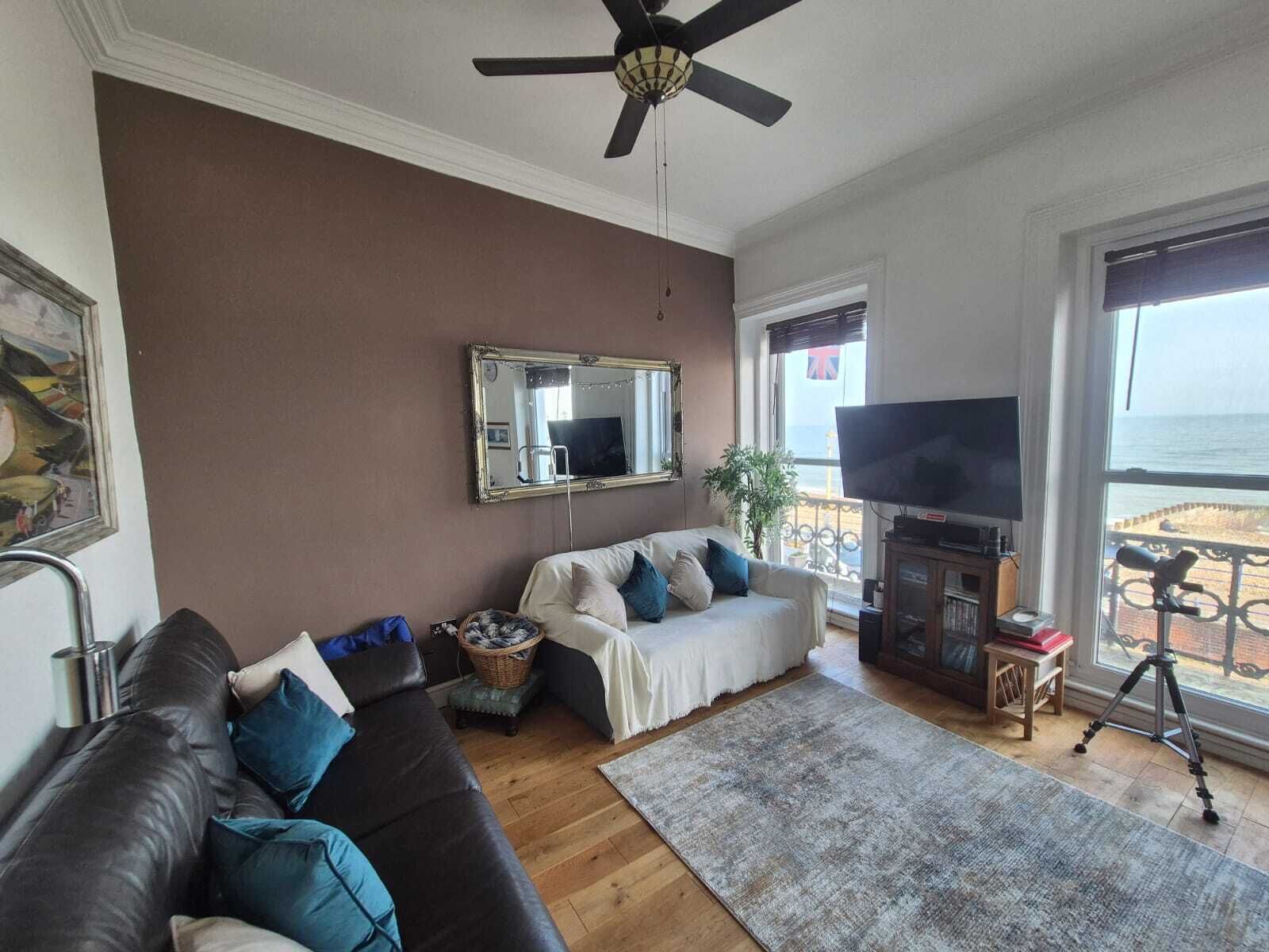 A living room with a brown wall, a ceiling fan, a large mirror, a sofa, a TV, and a view of the sea through large windows.