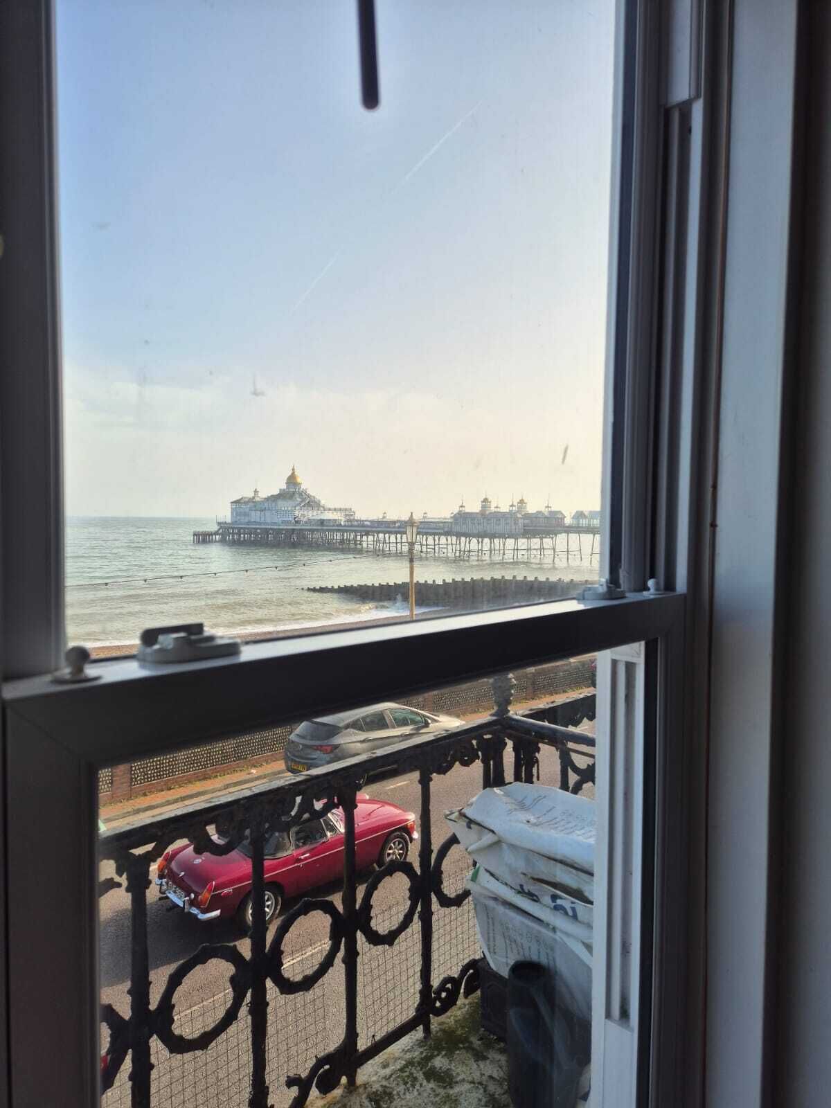 A view of a pier extending into the water with a building at the end, a balcony with a railing, and a red car parked on the street.