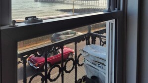 Vista de un muelle que se adentra en el agua, con un edificio al final, un balcón con barandilla y un coche rojo estacionado en la calle.