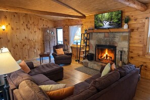 Ein gemütliches Blockhaus-Interieur mit Kamin aus Stein, Holzwänden, einer bequemen Couch und einem Fernseher, der über dem Kamin angebracht ist.