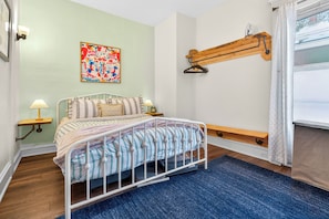 A bedroom with a bed, a wooden shelf, a window with curtains, and a colorful wall decoration.