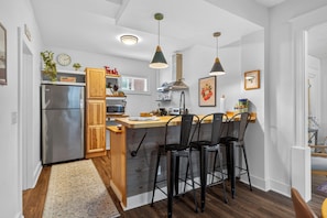 A kitchen with a stainless steel refrigerator, wooden cabinets, a wooden island with bar stools, pendant lights, and a clock on the wall.