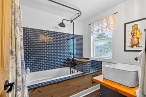 A bathroom with a bathtub, a shower, a window with a view, and a framed picture on the wall.