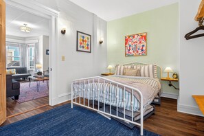 A bedroom with a bed, a nightstand, a lamp, a colorful painting, and a view of a living room with a sofa and a rug.