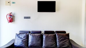 Un sofá de cuero oscuro con cuatro cojines, un extintor, un televisor y un interruptor de luz en la pared.