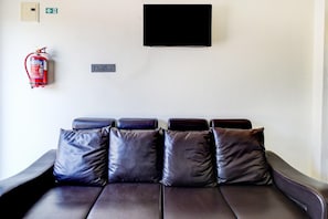 Un sofà de cuir fosc amb quatre coixins, un extintor, una televisió i un interruptor de llum fixat a la paret.