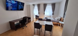Quarto de hotel pequeno com cama, escrivaninha, cadeira, televisão e janela com cortinas.