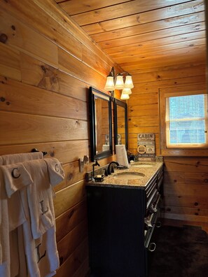 Une salle de bain de style rustique, avec des murs en bois, un grand miroir, un lavabo doté d’un plan de travail en granit et une fenêtre avec des stores.