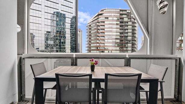 Un comedor con una mesa y sillas, vista a edificios modernos a través de una ventana, y una pequeña planta en maceta sobre la mesa.
