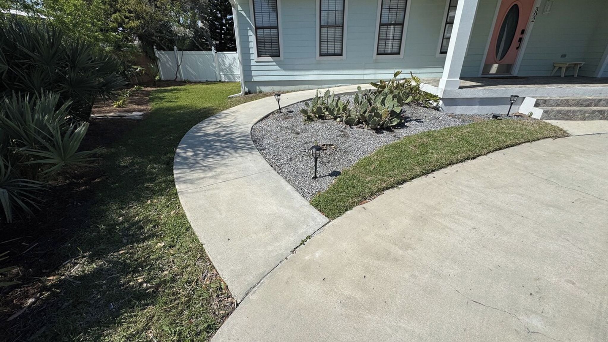A residential driveway with a concrete path, a small garden area with cacti, and a white house with a porch.