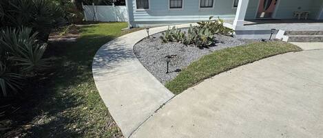 A residential driveway with a concrete path, a small garden area with cacti, and a white house with a porch.