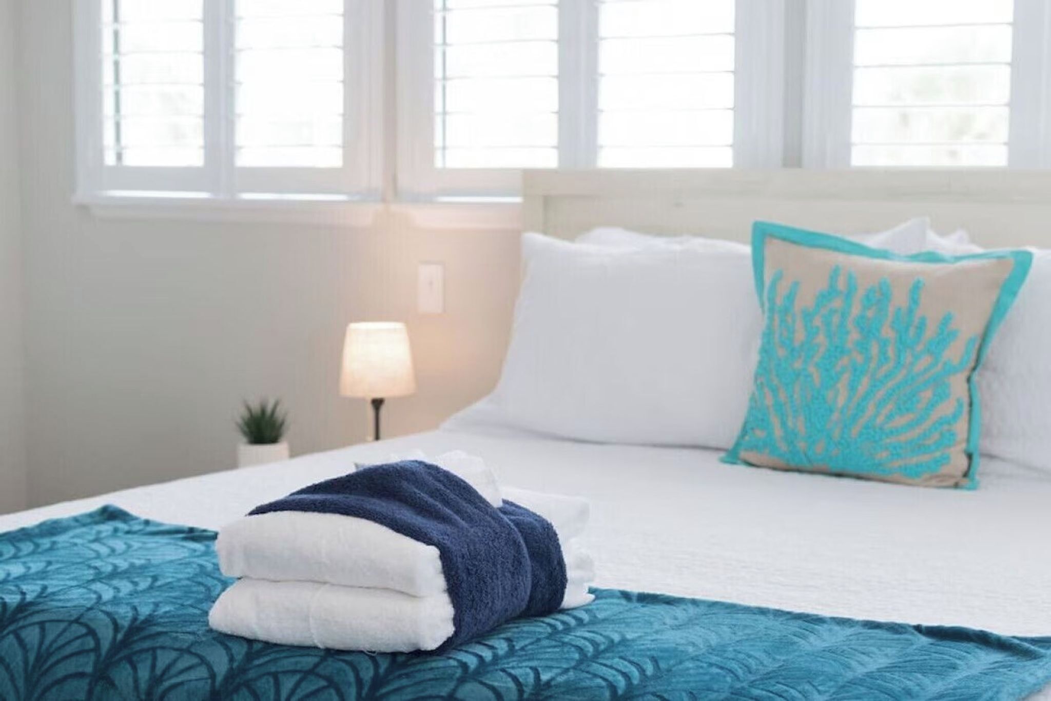 Een net opgemaakt bed met wit en blauw beddengoed, een blauwgroene plaid en een koraalgekleurd kussen.