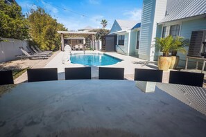 Una piscina con sillones de descanso, un porche cubierto y una casa de fachada azul.
