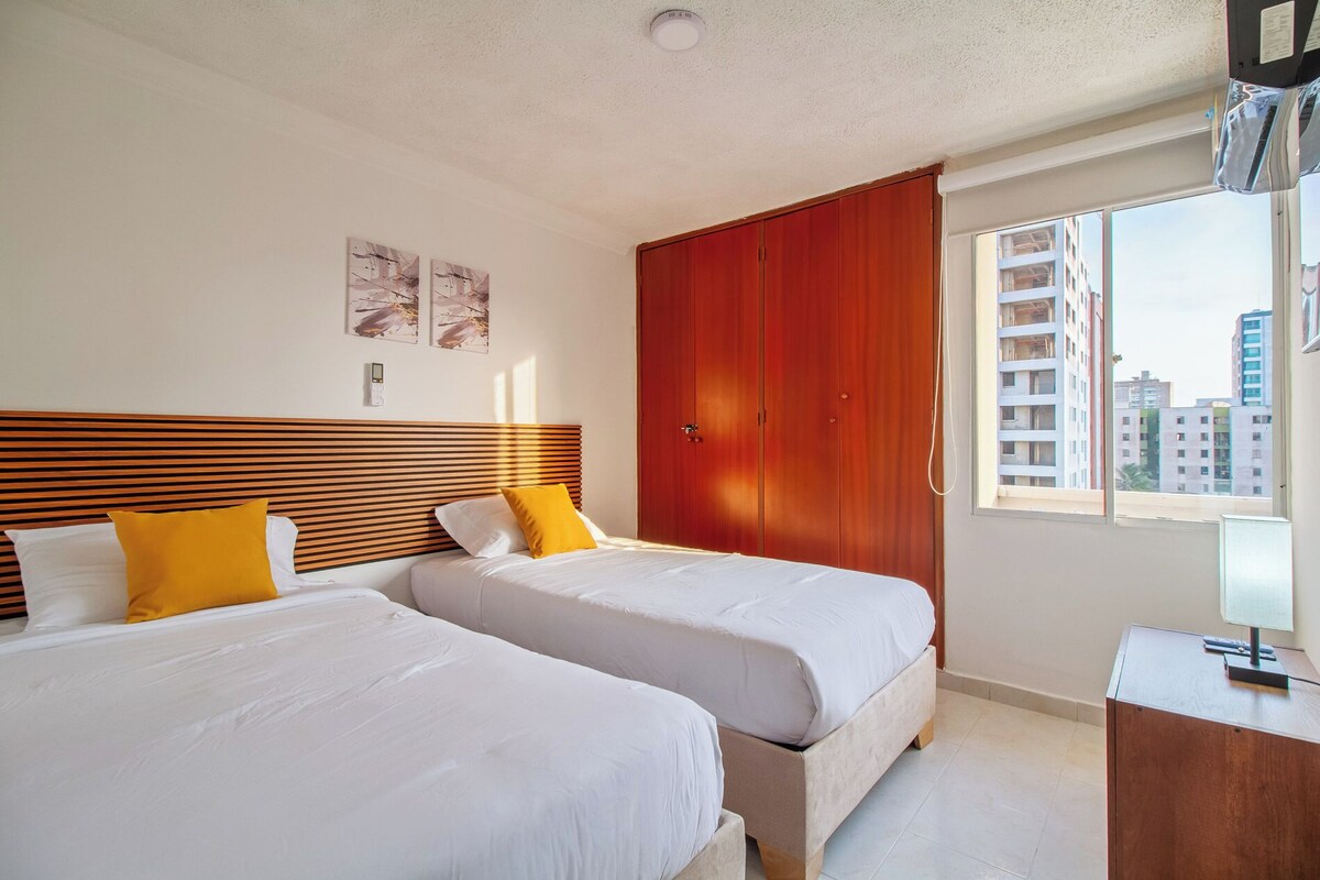 A hotel room with two beds, a wooden headboard, a wardrobe, a desk, and a window with a city view.