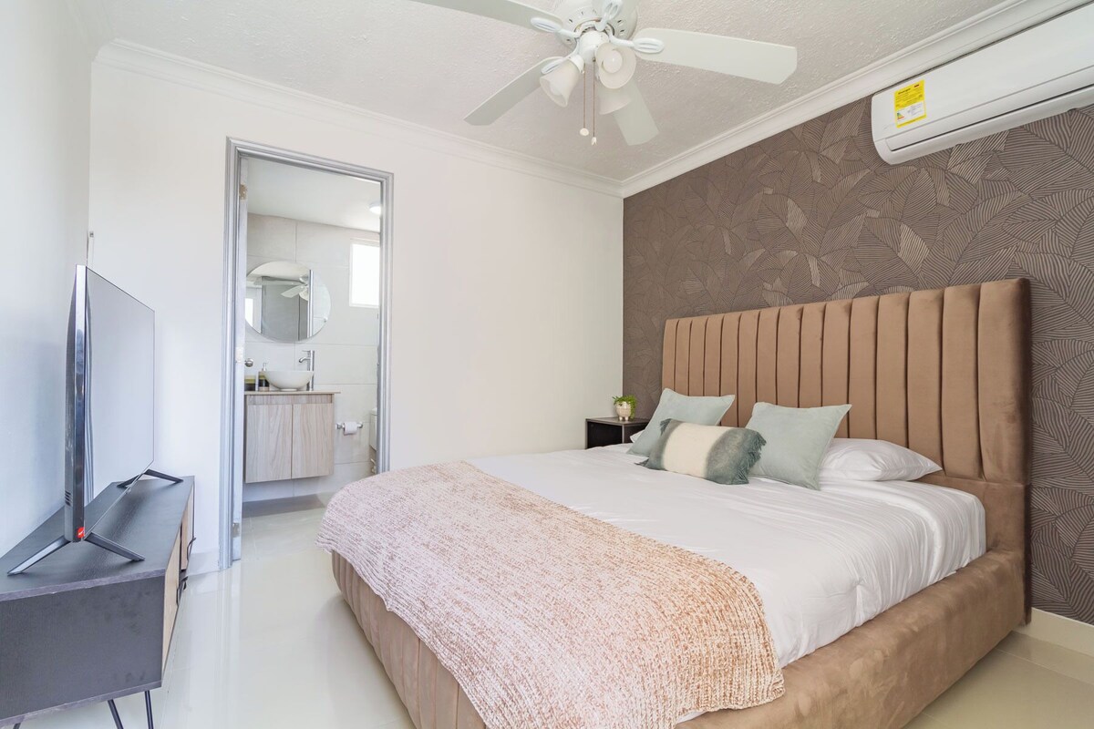 A bedroom with a bed, a TV, a ceiling fan, and a bathroom visible through an open door.
