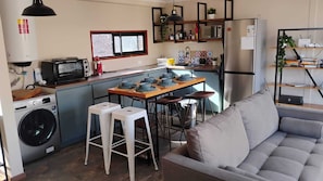 A compact kitchen with a washing machine, a microwave, and a refrigerator. There is a dining area with stools and a table, and a grey sofa in the living area. - Gorgeous 3-bedroom farmhouse in charming San Esteban awaits you (San Esteban)