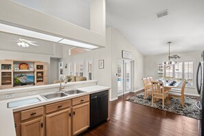 A modern kitchen with a dishwasher, double sink, and wooden cabinets. There is a dining area with a table and chairs, and a kitchen island with a sink. - Loma Vista Escape - 4BR Pool Home Near Disney, Golf & Shopping (Davenport)