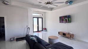 A spacious living room with a large black sofa, a ceiling fan, a TV mounted on the wall, and a wooden cabinet. - Spacious 4-bedroom tranquil escape Lehigh Acres Near the Forum (Lehigh Acres)