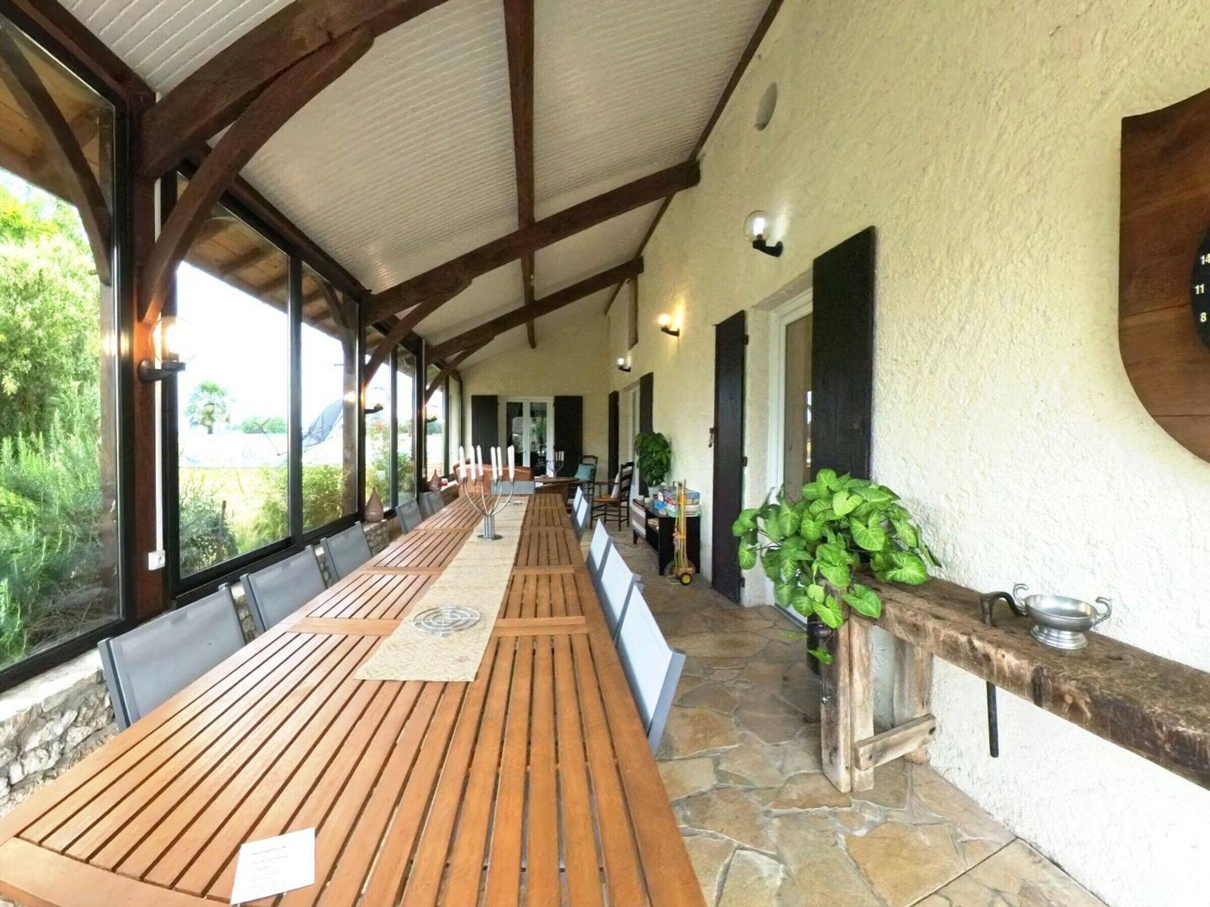 A covered outdoor dining area with wooden tables and chairs, stone flooring, and large windows offering a view of greenery.