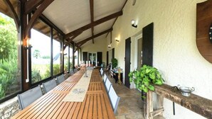A covered outdoor dining area with wooden tables and chairs, stone flooring, and large windows offering a view of greenery. - Cottage with private pool - 4 guests (Vanxains)