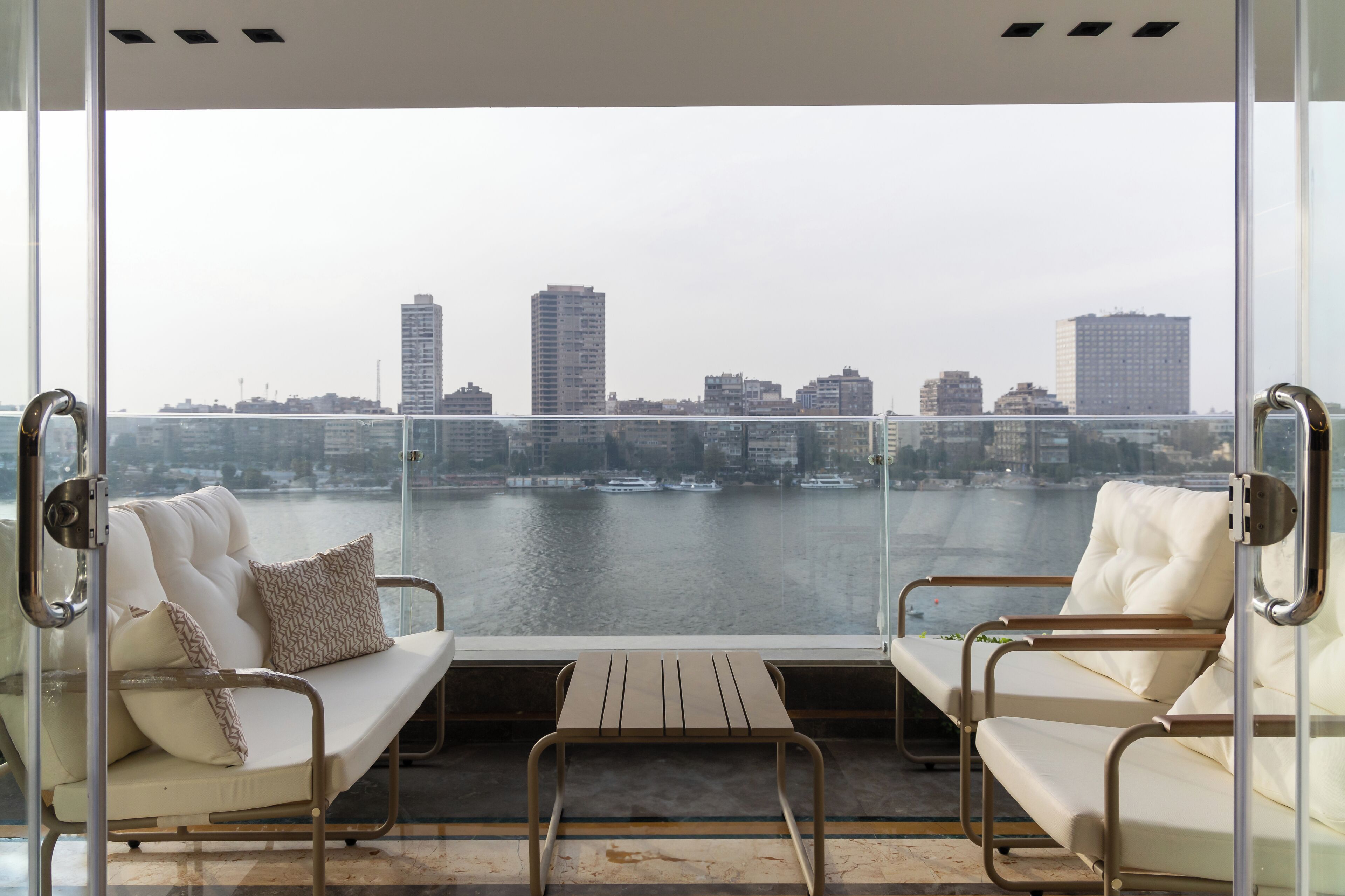 A balcony with two white cushioned chairs and a small table, overlooking a cityscape and water.