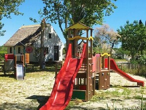 Un terrain de jeux avec un toboggan et des structures d’escalade, un bâtiment blanc avec un toit brun, et des arbres en arrière-plan.