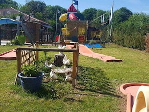 Un terrain de jeux équipé de toboggans et de structures d’escalade, entouré d’une pelouse, d’une clôture en bois et d’une petite table agrémentée d’objets décoratifs.