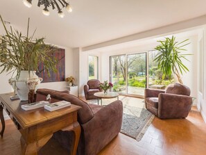 Ein modernes Wohnzimmer mit einem Holztisch, zwei braunen Sesseln, einer großen Vase mit Trockenblumen und Blick auf einen Garten durch große Schiebeglastüren.